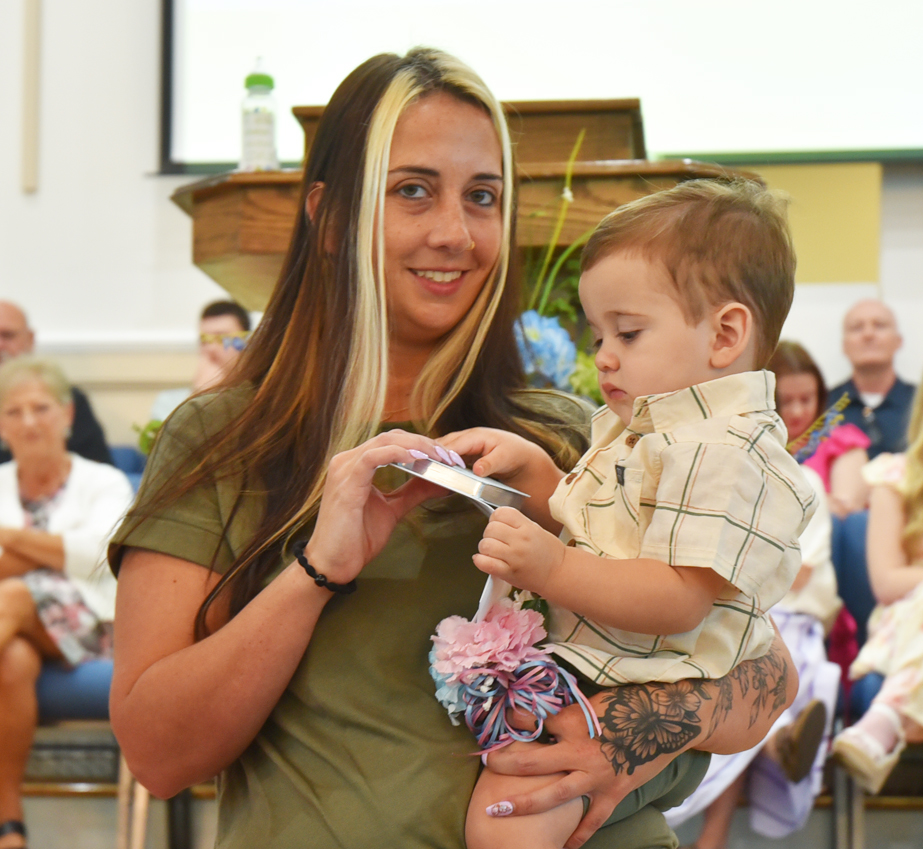 Woman holding a young child in an indoor setting, both looking at a piece of paper, with people seated in the blurred background.