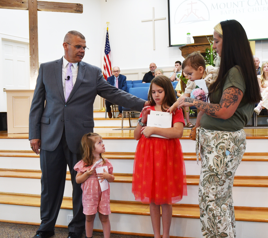 A man in a suit stands with a woman and three children on church steps, holding papers during a ceremony. Congregation and cross visible in the background.