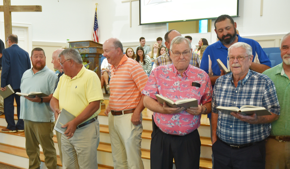 A group of men holding hymnals stand at the front of a church, smiling, with a choir and congregation seated in the background.