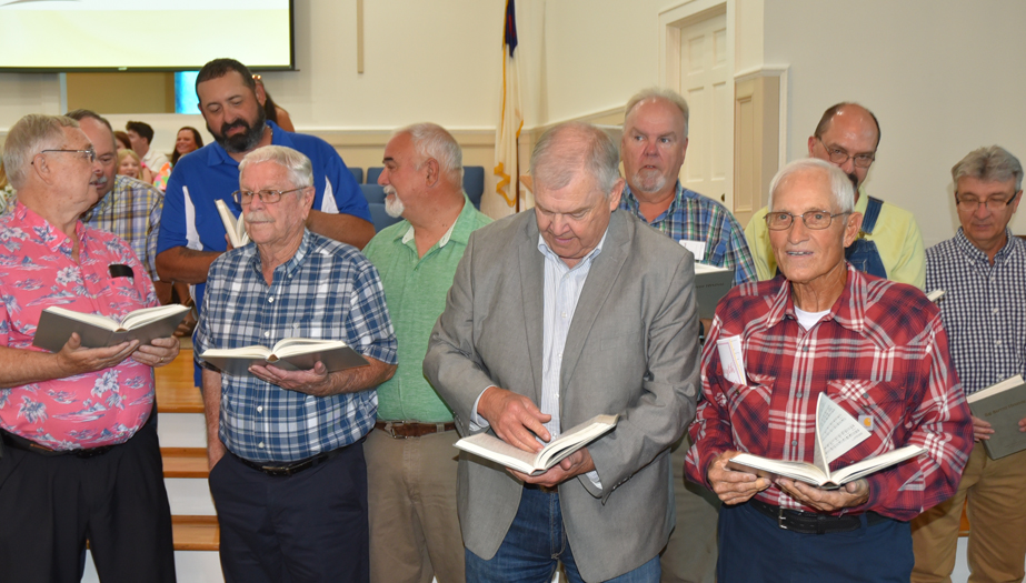A group of older men standing together in a church, holding and reading from hymnals, appearing to sing or prepare for a service.