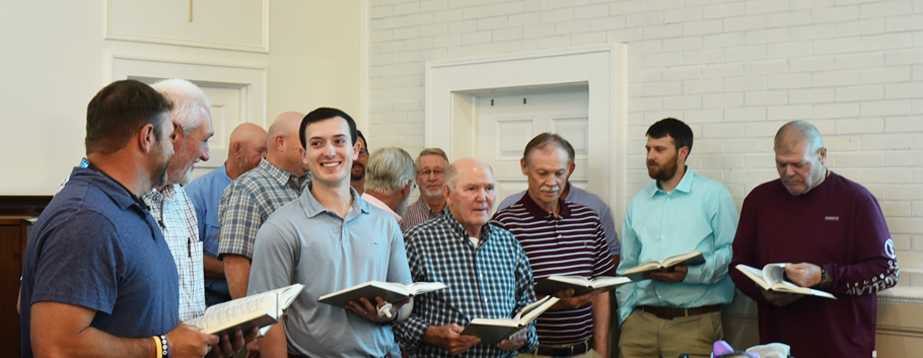 A group of men standing indoors, holding hymn books and singing together, with a white brick wall in the background.