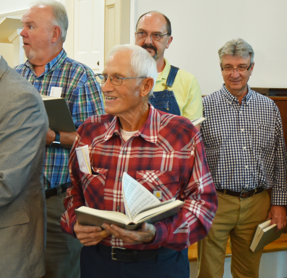 A group of older men stand together indoors, each holding a book and appearing to sing or participate in an event, dressed in casual plaid shirts.