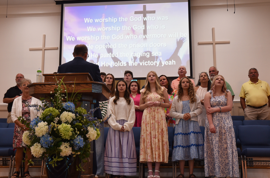 A church choir stands on stage singing in front of a projected worship song with lyrics, floral arrangement, and crosses on the walls.
