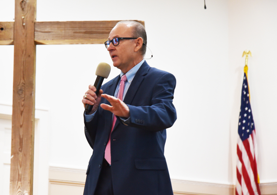 A man in a suit speaks into a microphone near a wooden cross and an American flag in a church or meeting hall.