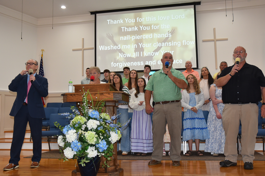 A church choir sings on stage with microphones, floral arrangement in front, song lyrics projected on screen behind, and congregation dressed in semi-formal attire.