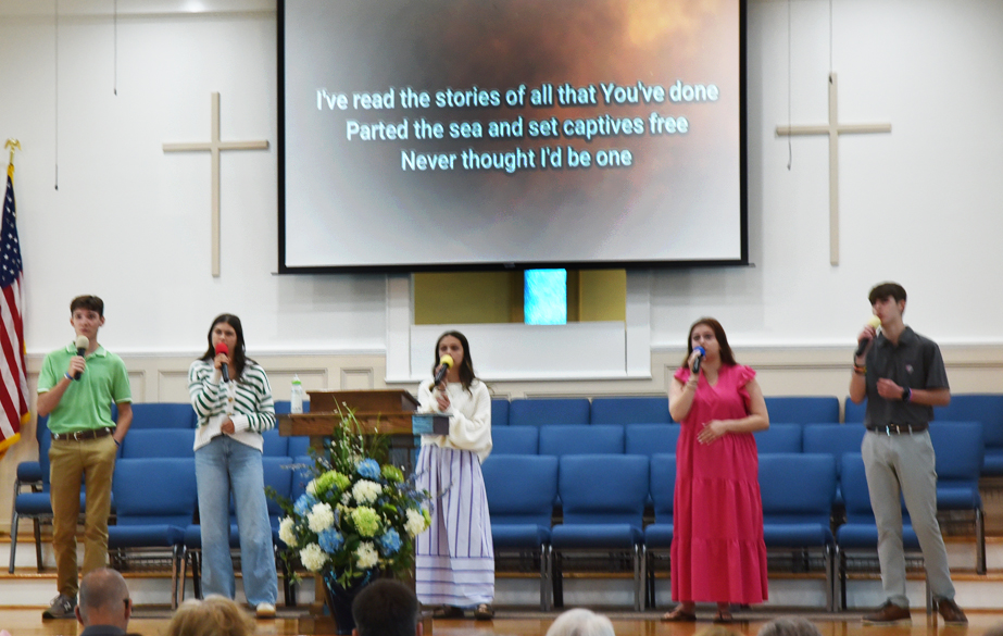 Five people singing on a church stage with microphones, a flower arrangement, and song lyrics projected on a screen above them.