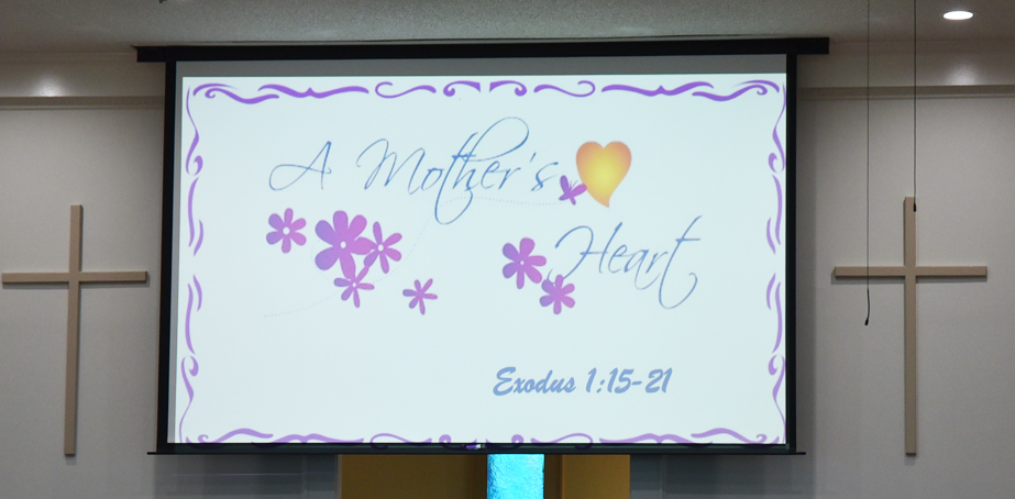 A church projector screen displays "A Mother's Heart" with purple flowers, heart graphic, and Bible reference Exodus 1:15-21, flanked by two crosses on the wall.