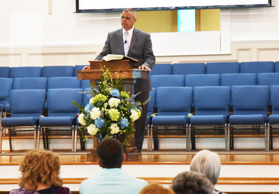 A man in a suit speaks at a podium in a church, with blue chairs and a floral arrangement, while people listen from the audience.
