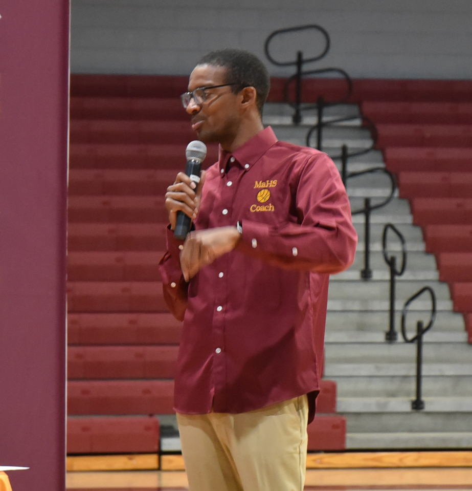 A man wearing glasses and a maroon shirt labeled "MaHS Coach" speaks into a microphone in a gymnasium with red bleachers.