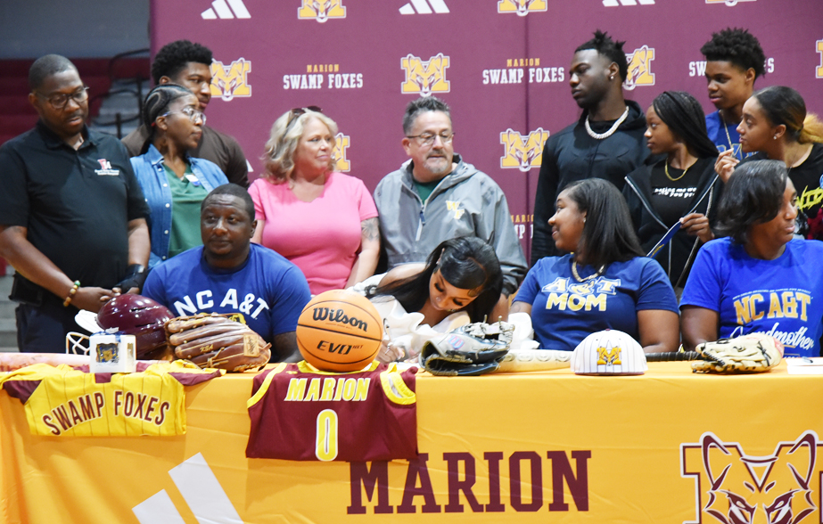 Student signs letter of intent at Marion Swamp Foxes event, surrounded by family, friends, and sports memorabilia including basketball, football, and jersey.