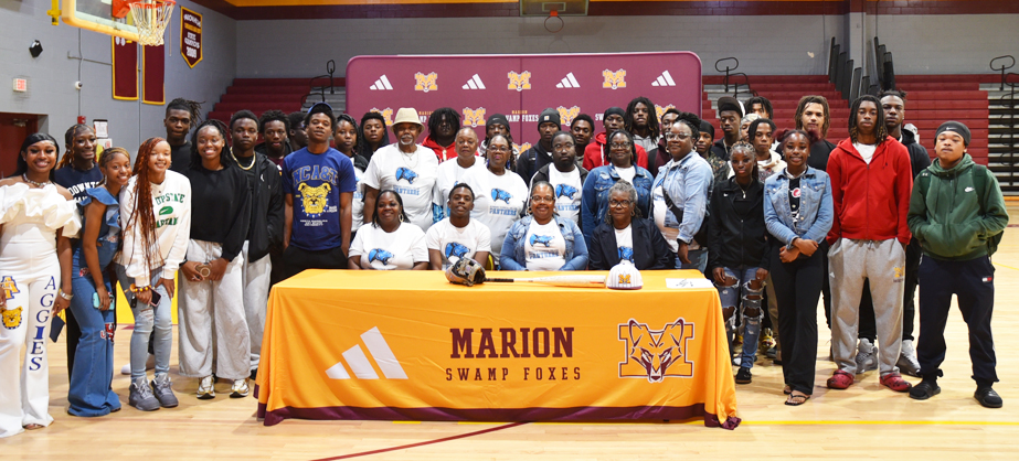 A large group of students and adults stand behind a signing table with a Marion Swamp Foxes banner in a school gymnasium.