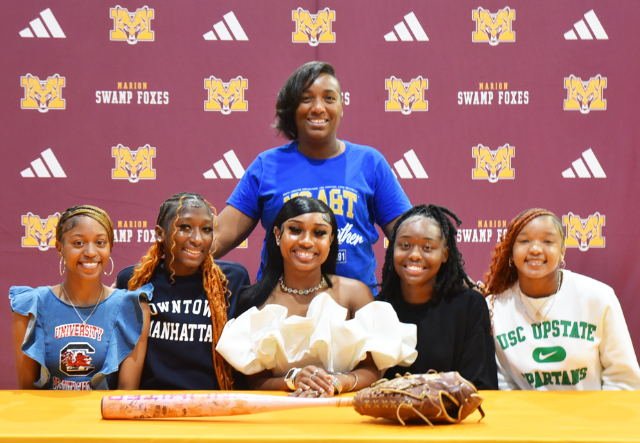 Six people pose and smile at a table with a bat and glove, in front of a Marion Swamp Foxes backdrop.