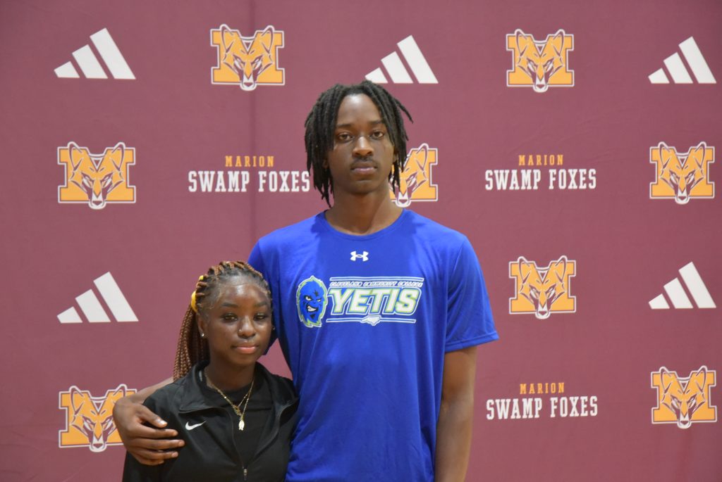 Two people pose together in front of a Marion Swamp Foxes backdrop, one wearing a blue Yetis shirt and the other dressed in black sportswear.