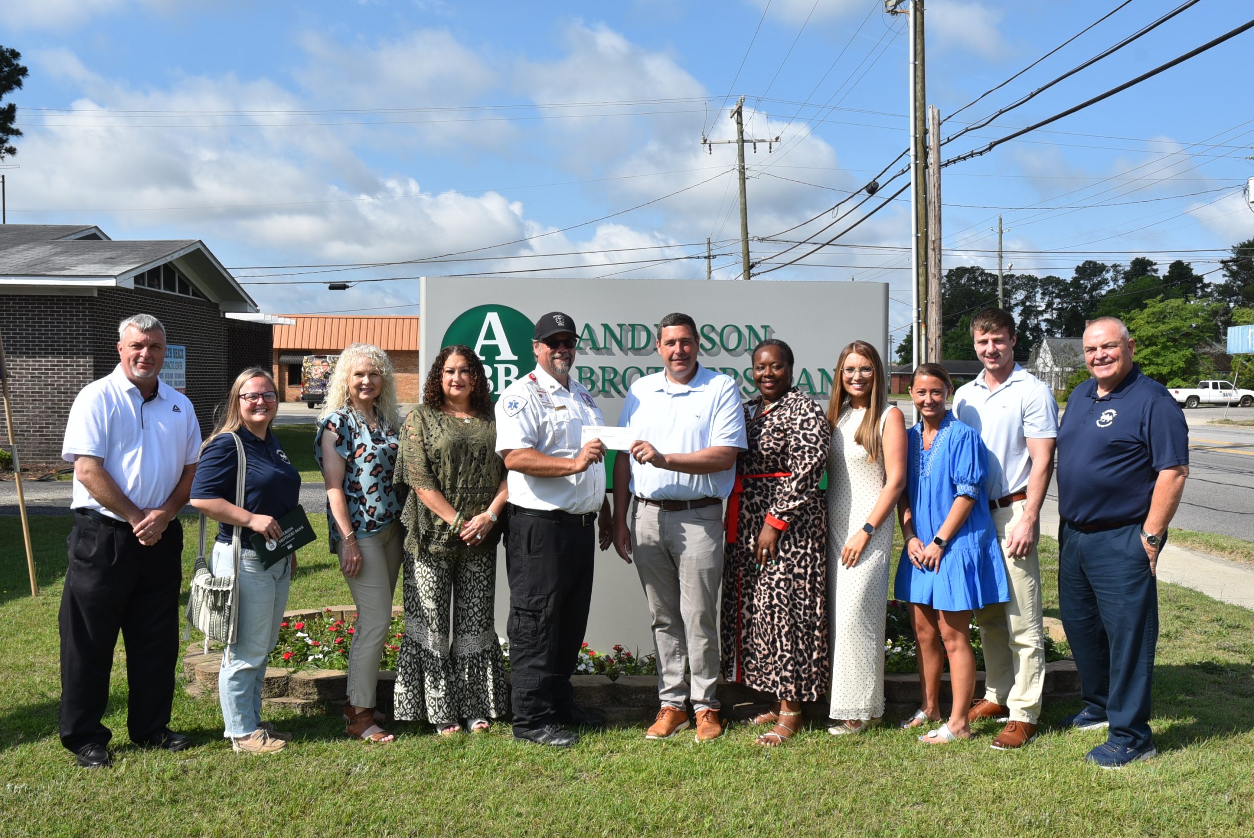A group of people standing in front of the Anderson Brothers Bank sign, smiling for a photo on a sunny day.