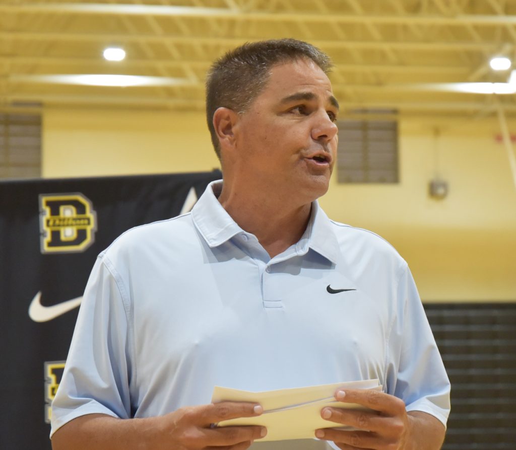 Man wearing a light blue Nike polo shirt speaking indoors, holding papers, with a black and yellow banner in the background.