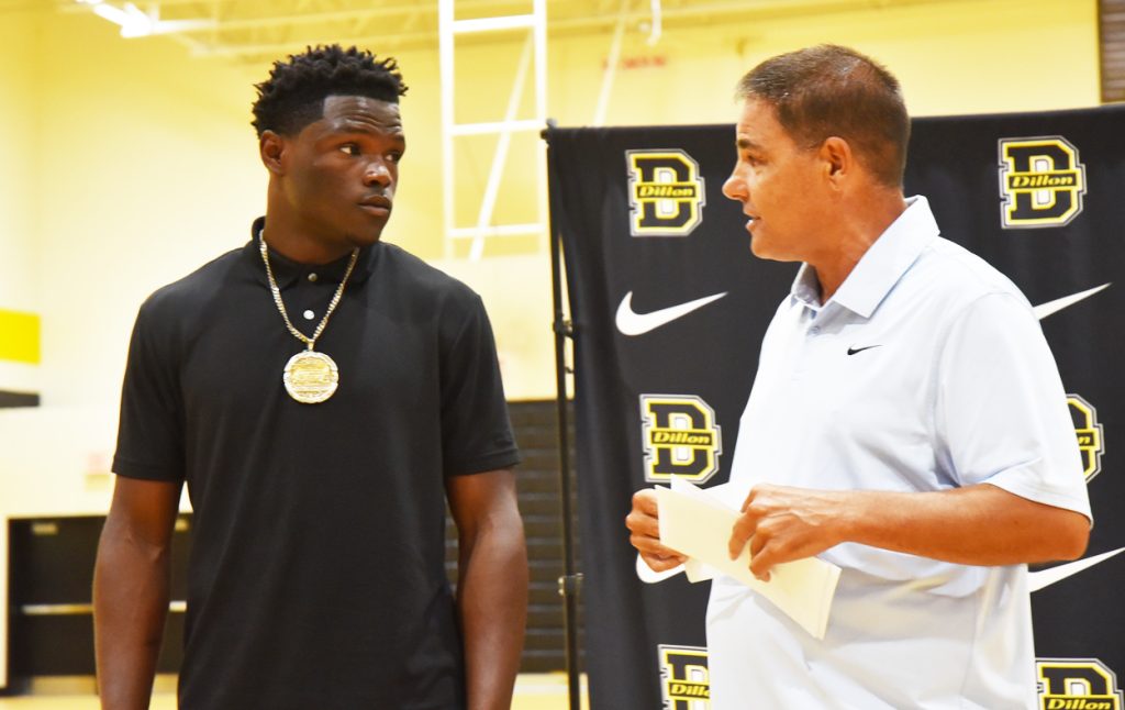 Two men stand in a gymnasium having a conversation, with one holding papers. A "Dillon" logo and Nike symbol are displayed on the background banner.