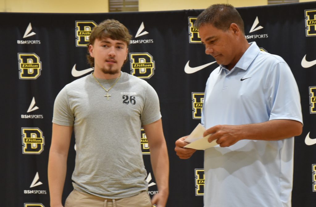 Two men standing indoors in front of a black backdrop with Dillon High School and Nike logos, one holding papers.