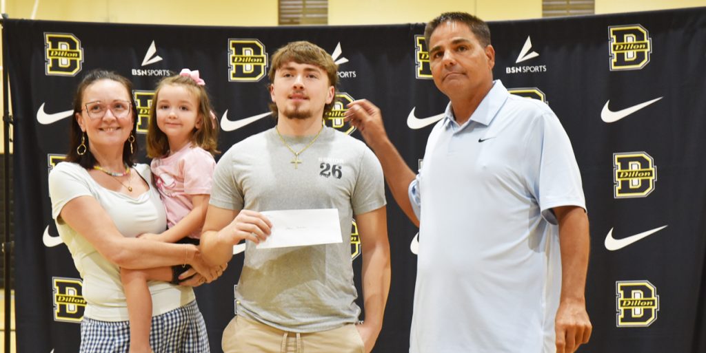 Four people standing together, one holding an envelope, in front of a Dillon High School backdrop with Nike and BSN Sports logos.