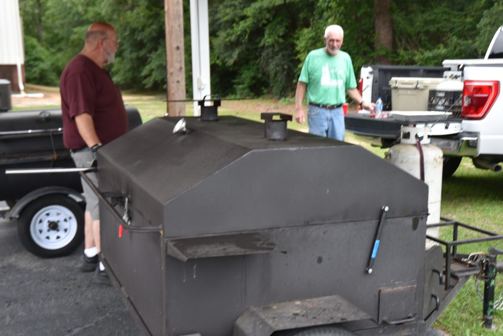 Two men stand beside a large black outdoor barbecue smoker under a canopy, with trees and grassy area in the background.