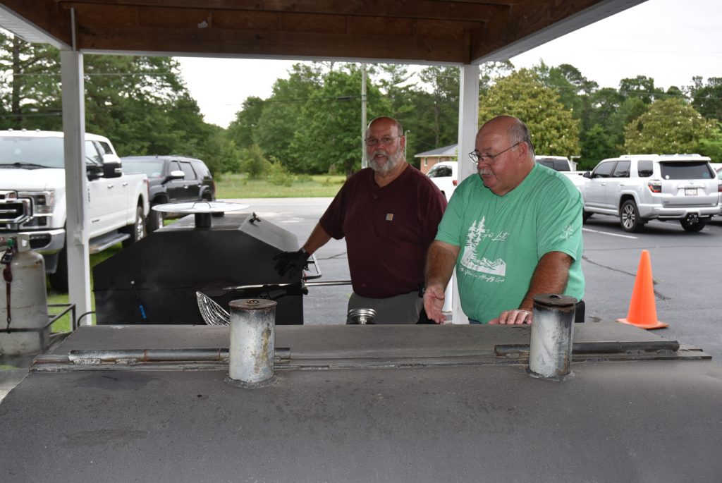 Two men stand behind a grill under a shelter, with parked vehicles and trees visible in the background.