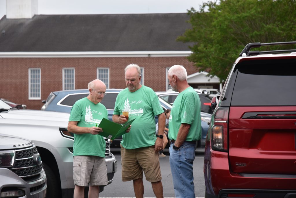 Three older men in matching green shirts stand talking between parked cars outside a brick building, one holding a folder or clipboard.