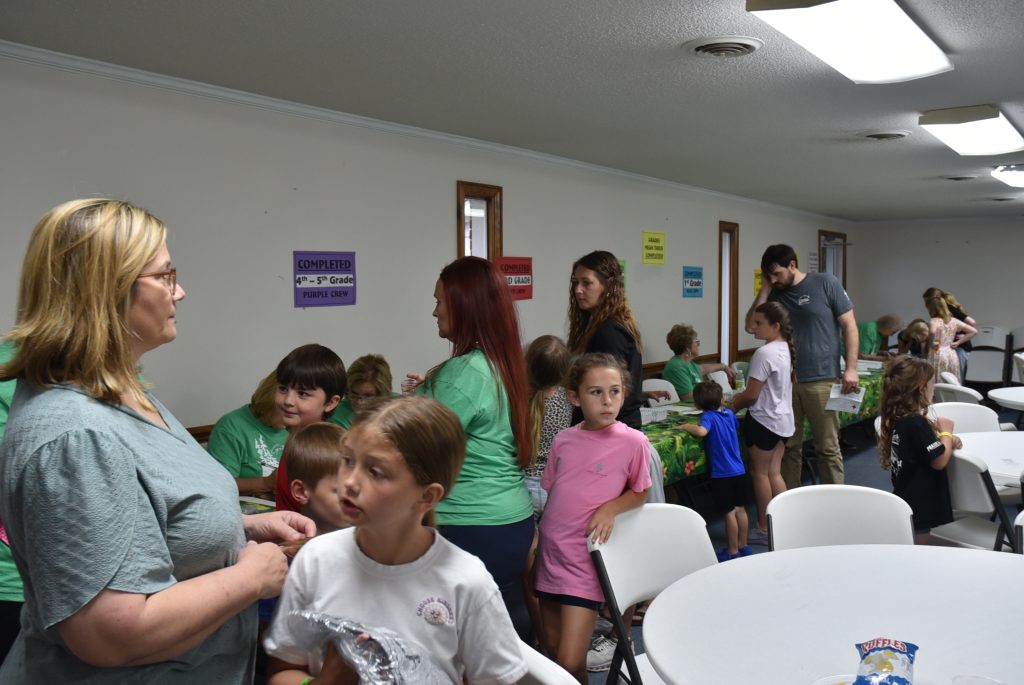 Children and adults gather in a brightly lit room, engaging in activities around tables. Colorful posters decorate the walls.