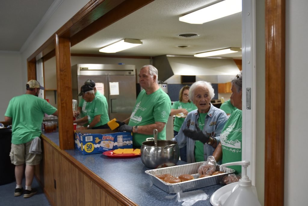 Volunteers wearing green shirts serve food in a cafeteria kitchen, preparing meals for a community event or charity service.