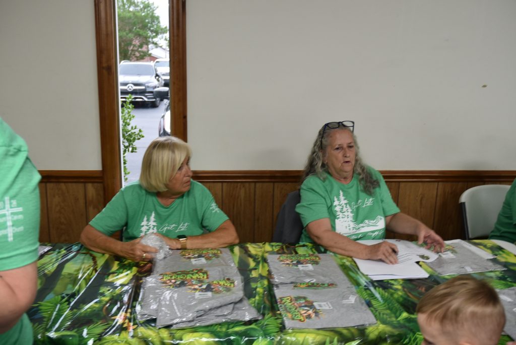 Two women in green shirts sit at a table with papers and t-shirts, appearing to assist with event registration indoors.