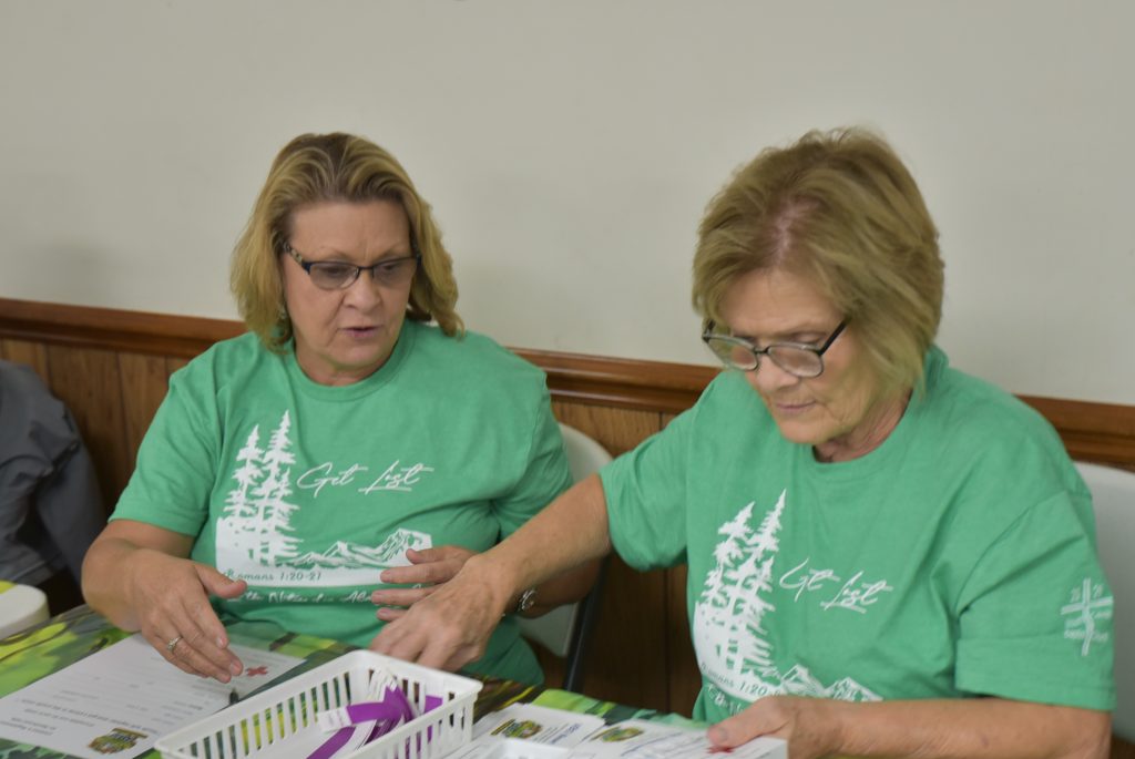 Two women in matching green shirts sit at a table, sorting papers together, with a white basket holding supplies in front of them.