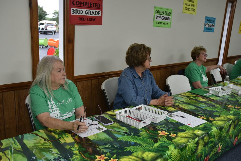 Three women sitting at a table covered with a green patterned cloth, taking notes, with colorful signs on the wall behind them.
