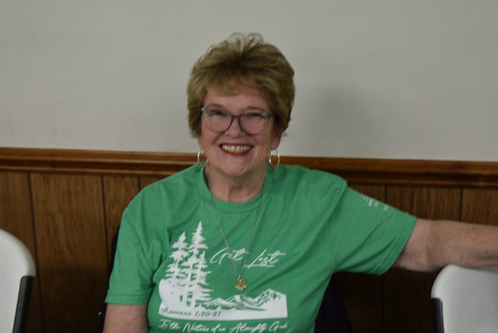 Smiling older woman with short hair and glasses, wearing a green graphic T-shirt, sitting indoors against a plain wall and wooden panel.
