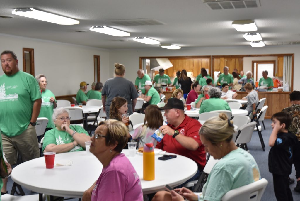 Large group of people sitting at round tables in a bright, indoor hall, wearing casual clothes and talking, with many dressed in green shirts.