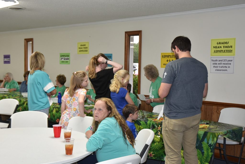 People gathered in a room, some sitting at tables with drinks, others standing and talking, colorful signs on the wall, indoors, casual atmosphere.