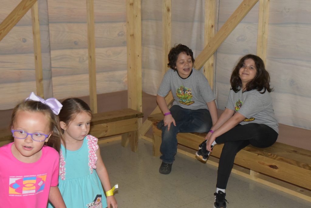 Three children are indoors; two are sitting on a wooden bench, one is standing. They are in a room with wooden walls.