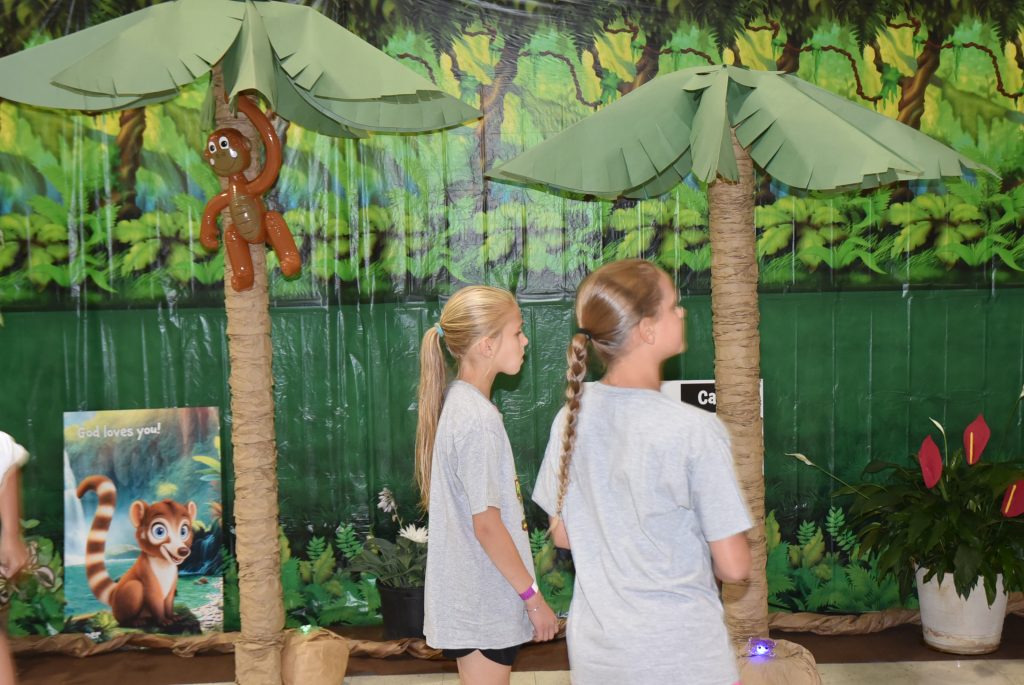 Two girls with braided hair walk past artificial palm trees and a jungle-themed backdrop, with a toy monkey hanging from a tree.