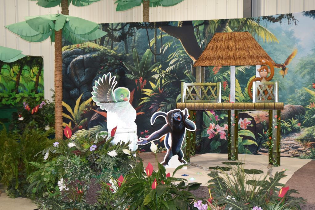 Jungle-themed display with cardboard cutouts of a bird and monkey, tropical plants, and a painted backdrop featuring huts and lush greenery.