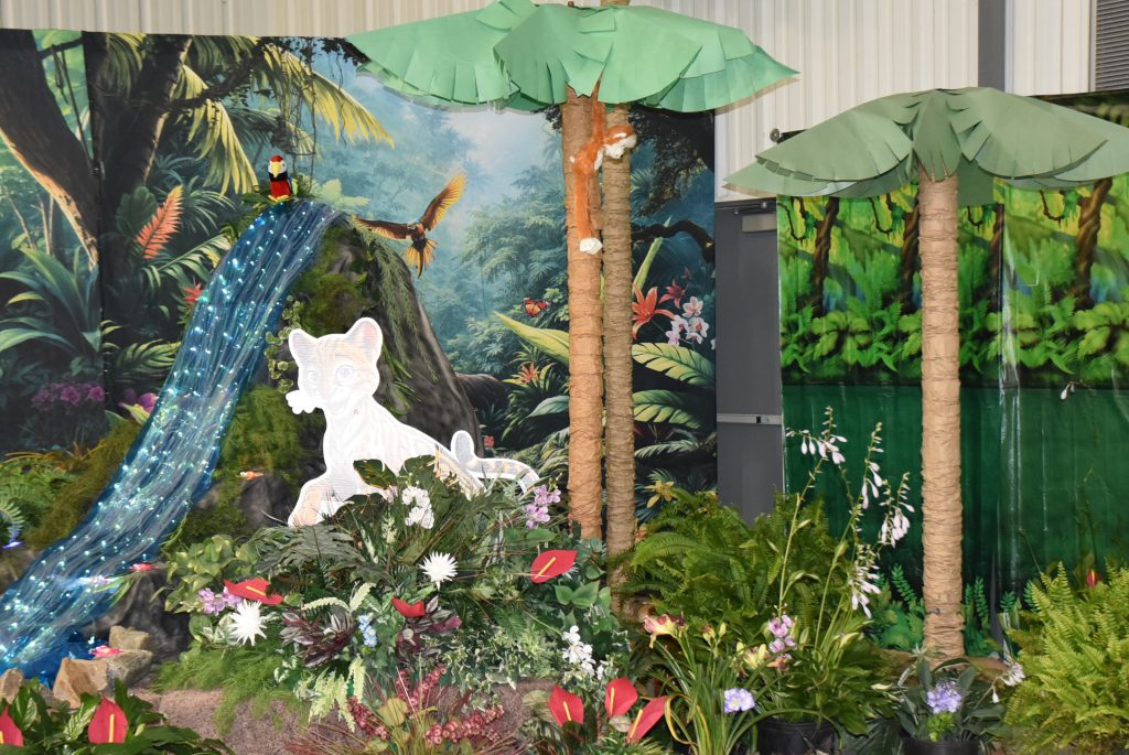 Jungle-themed display with artificial palm trees, lush plants, a white cutout of an animal, and a colorful rainforest backdrop featuring birds and a waterfall.