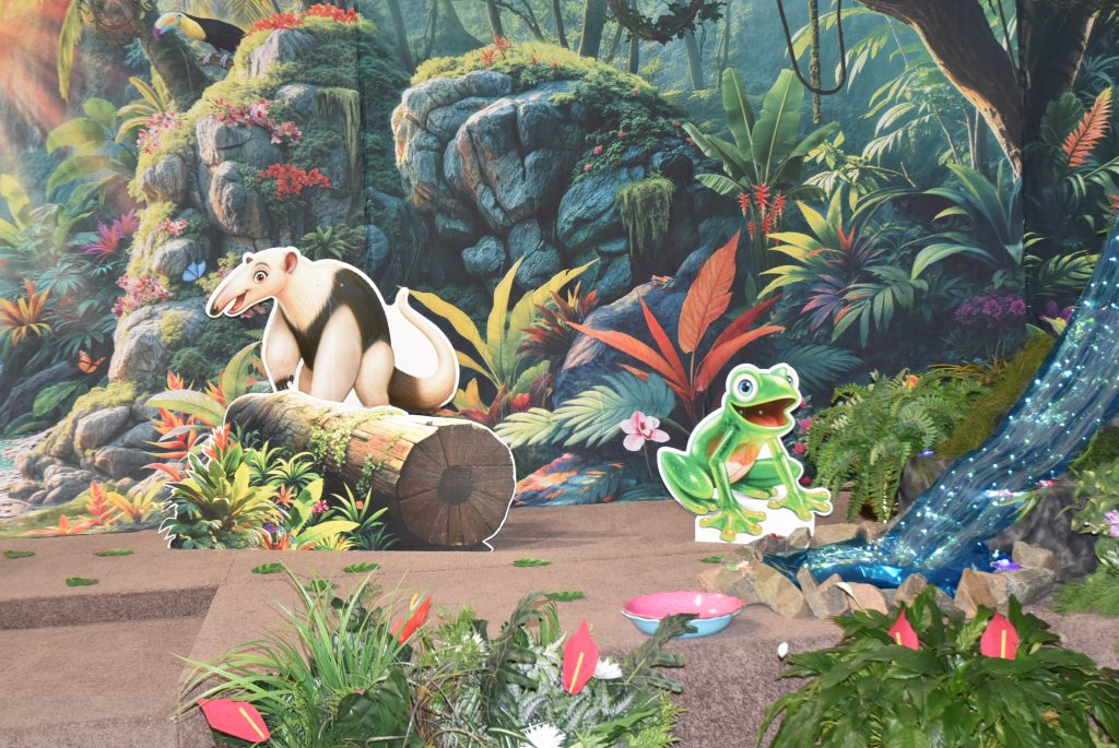 A colorful jungle scene with a tapir on a log and a cartoon frog near a small stream surrounded by lush plants and vibrant flowers.