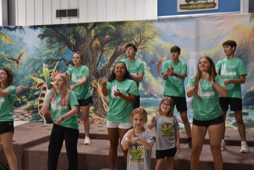 A group of children and teens in green shirts stand on a stage, smiling and gesturing in front of a colorful mural backdrop.