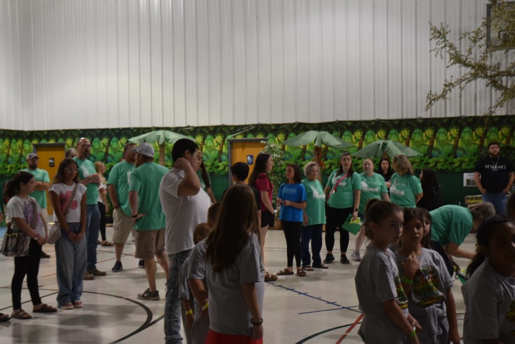 A group of people, including children and adults in green shirts, stand inside a gymnasium with a mural of green foliage on the back wall.