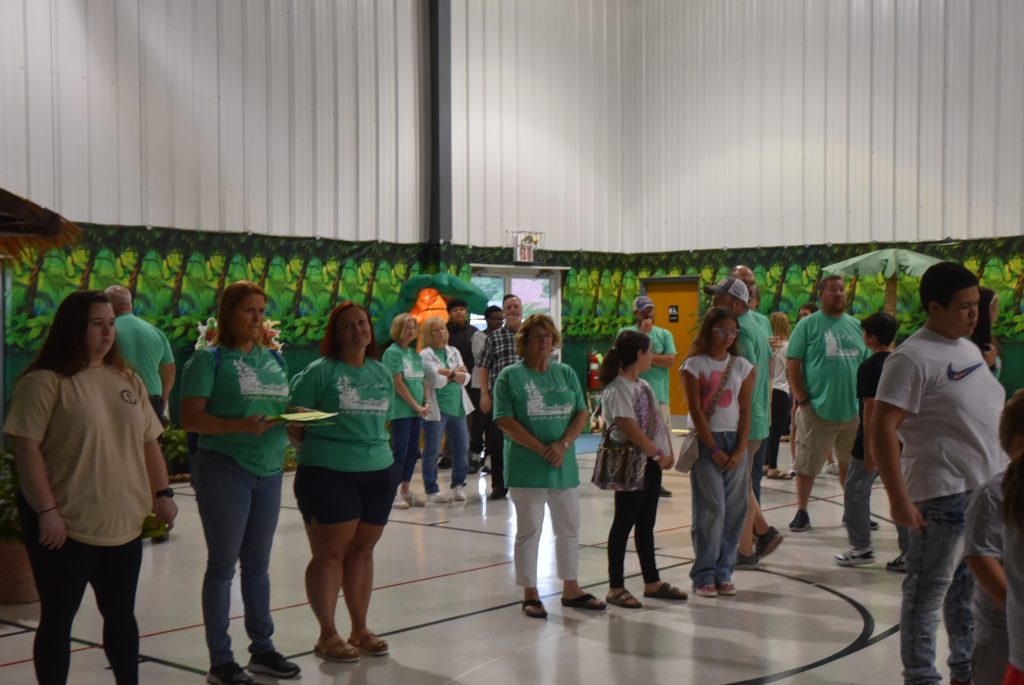 A group of people wearing green shirts stand inside a gymnasium, decorated with colorful wall art, participating in an indoor event or gathering.
