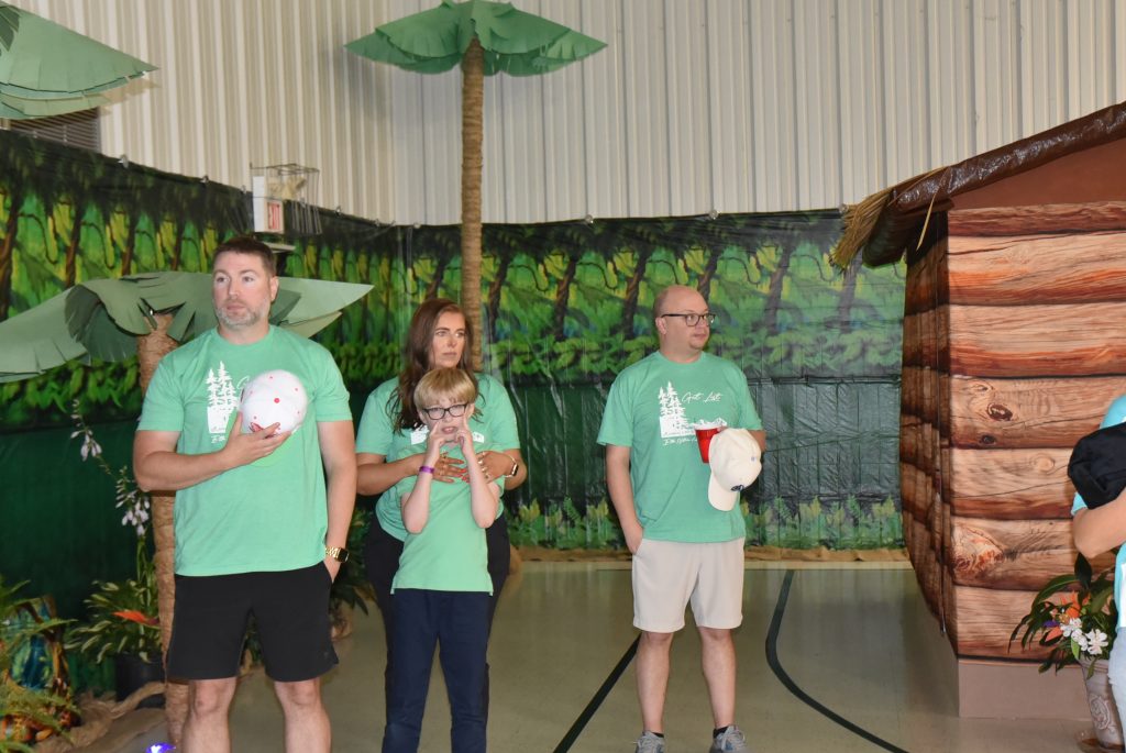 Three adults and one child wearing green shirts stand indoors with jungle-themed decorations and inflatable trees in the background.