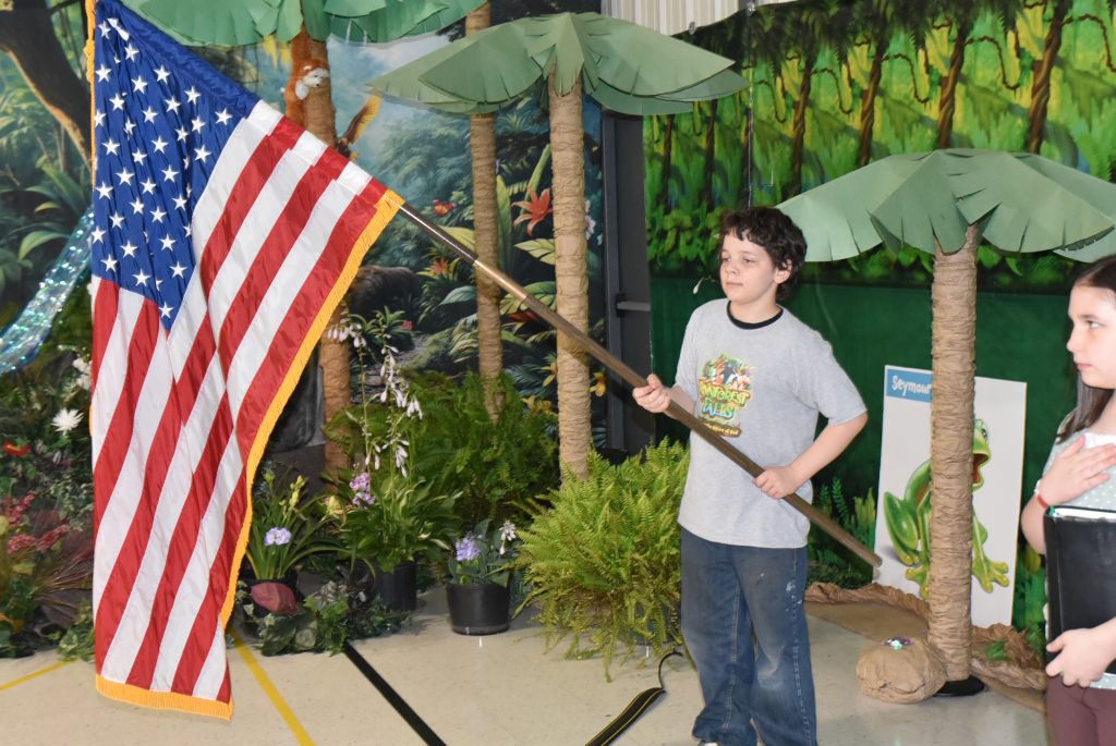 A young boy stands indoors holding an American flag, surrounded by green plants and a painted jungle backdrop.