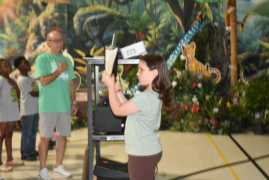 A young girl stands indoors holding a piece of paper, with a decorated jungle-themed background and an adult man watching nearby.