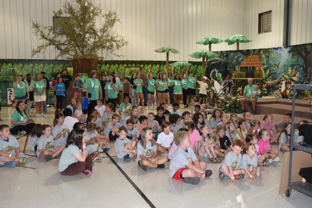 A group of children sit on the floor of a gymnasium, with adults standing behind them. The background is decorated with jungle-themed props and trees.