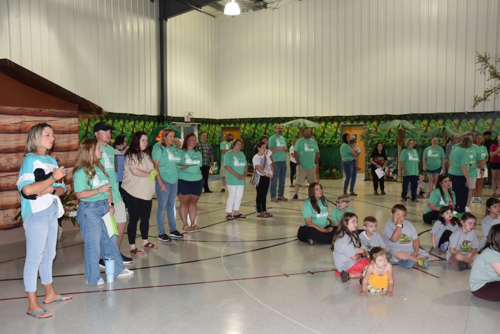 A group of adults and children gathered in a gymnasium, some standing and others sitting on the floor, wearing matching green shirts.