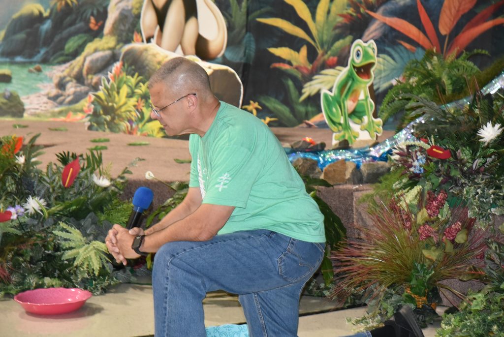 A man in a green shirt kneels in front of a jungle-themed backdrop with plants and a cartoon dinosaur.