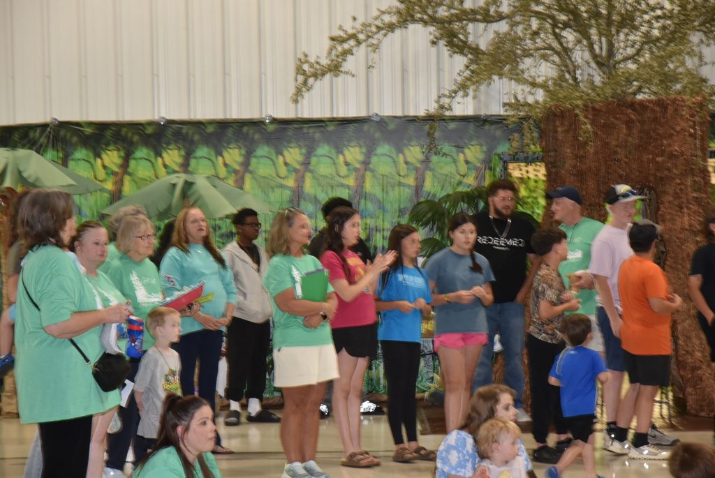 Adults and children stand indoors, some clapping and smiling, in front of a wall decorated with greenery.