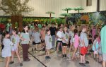 A large group of children and adults gather indoors, many wearing matching t-shirts. Artificial trees and jungle-themed decorations are visible in the background.