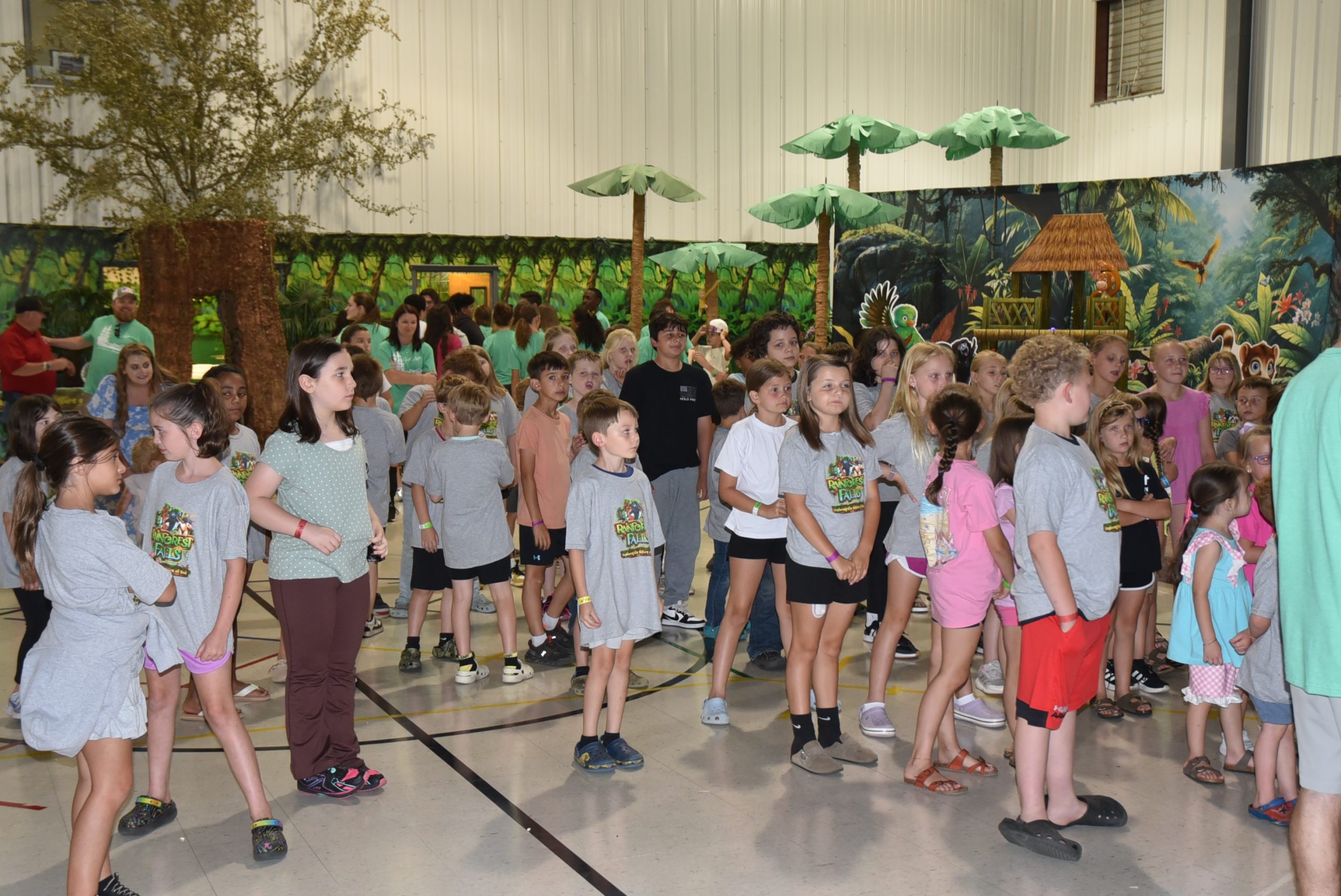 A large group of children and adults gather indoors, many wearing matching t-shirts. Artificial trees and jungle-themed decorations are visible in the background.